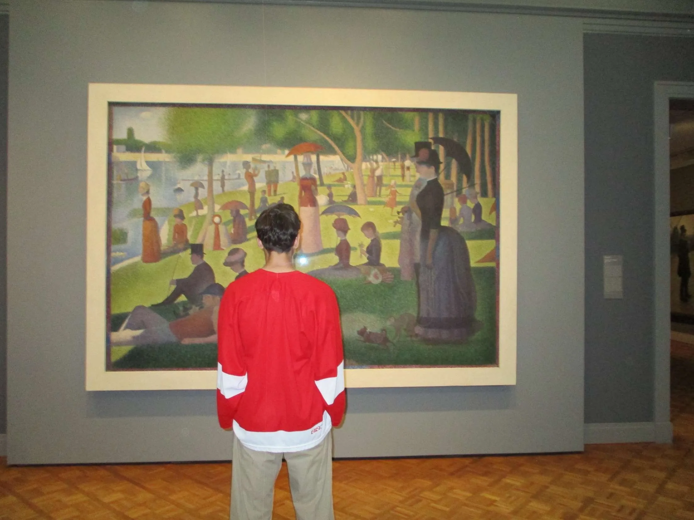 A visitor in a red hockey jersey viewing a pointillist park painting in a museum